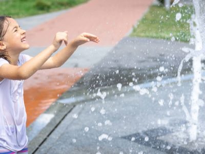 Happy little girl among the splashing water of the city fountain has fun and escapes from the heat.