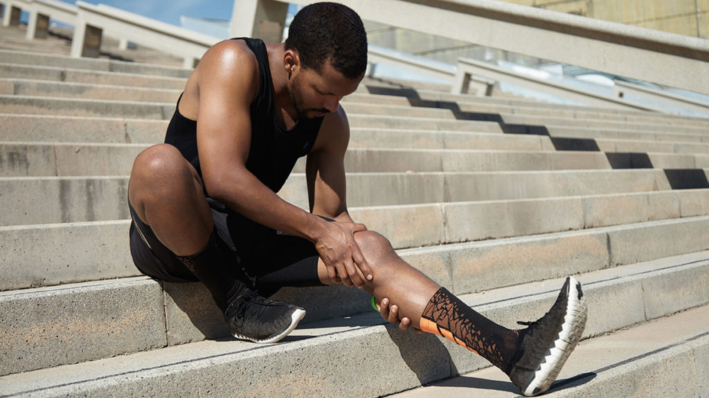 Handsome black runner with muscular athletic body holding his leg with both hands, feeling pain in knee or calf, massaging it, suffering from pain while sitting on steps of concrete stair