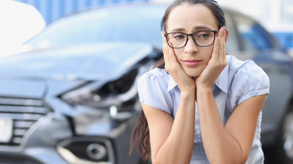 Portrait of a sad woman with totaled car in the background. Women is thinking about an auto injury chiropractor.