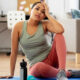 A tired woman in activewear sits on a yoga mat at home, holding her head to avoid strenuous physical exertion.
